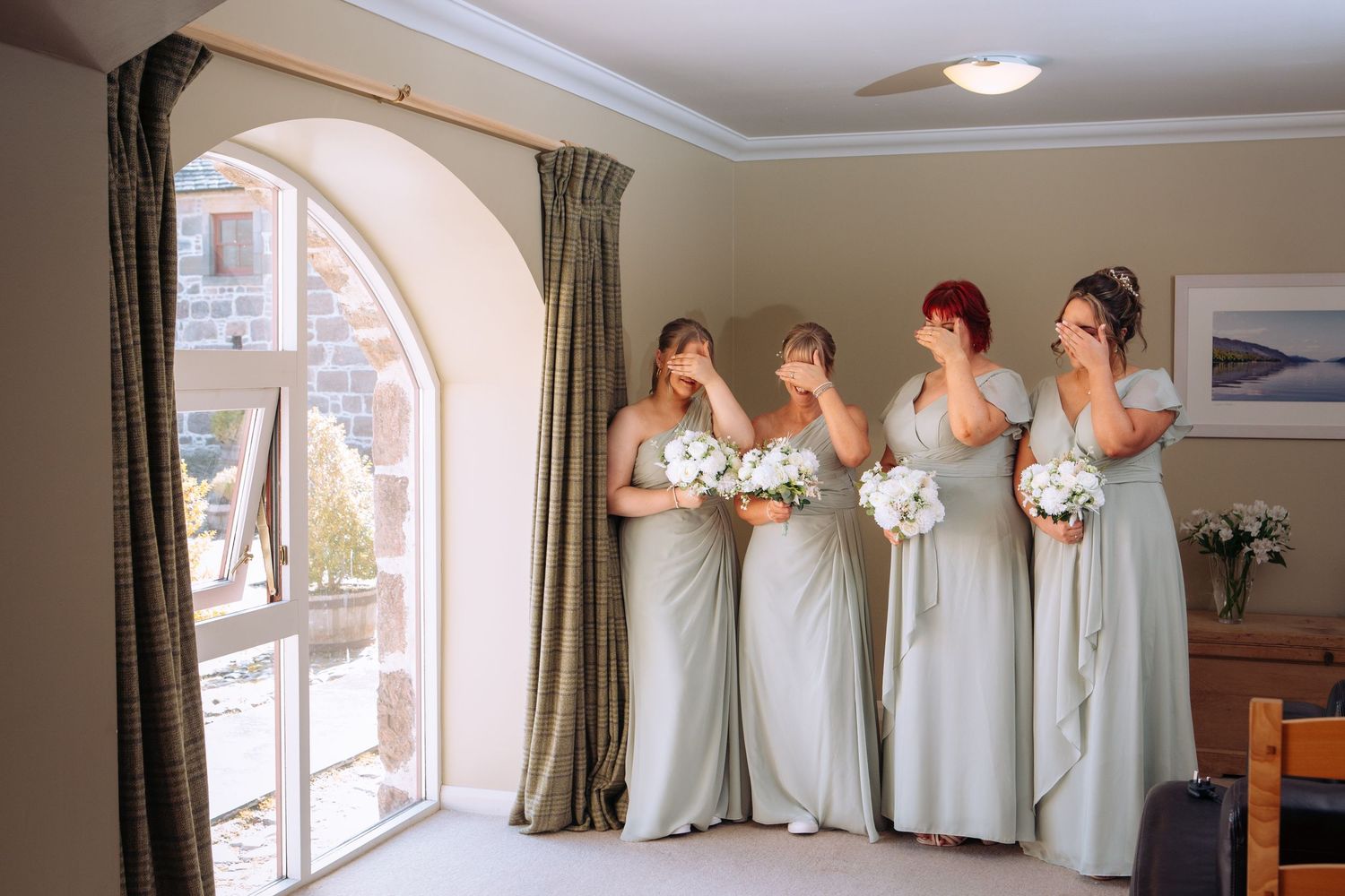Four bridesmaids in sage green gowns holding white bouquets, covering their faces with emotion during wedding preparations.