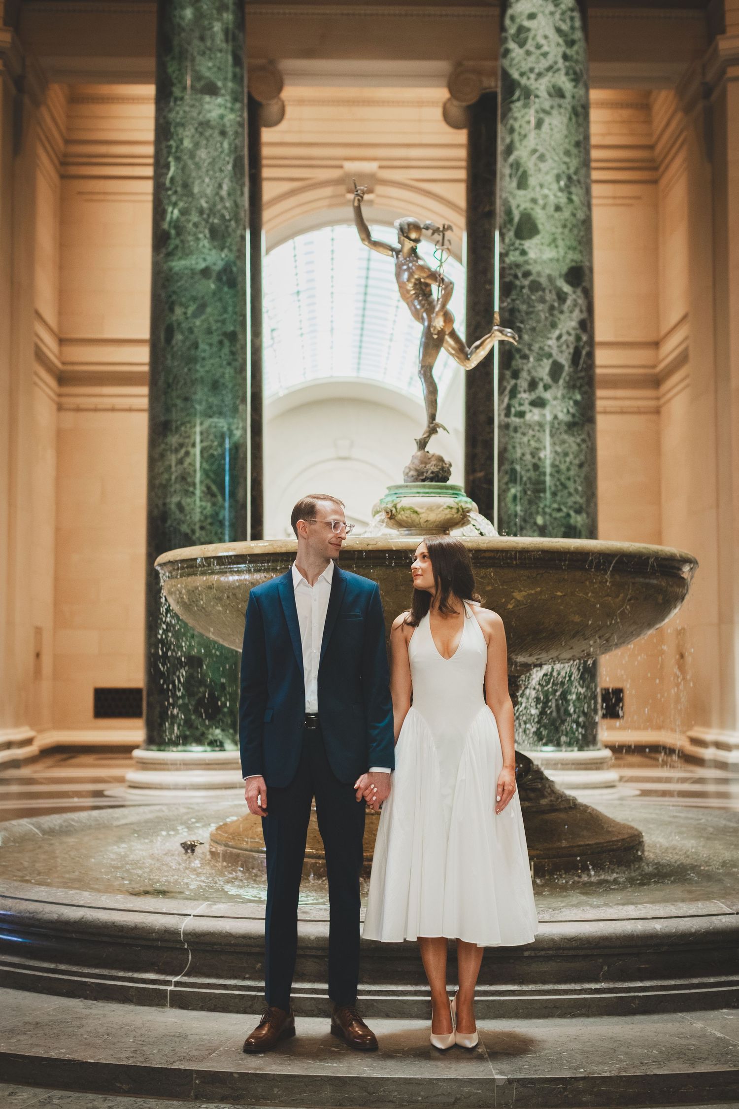 Couple in elegant wedding attire standing between draped curtains in a grand venue, sharing a romantic moment.