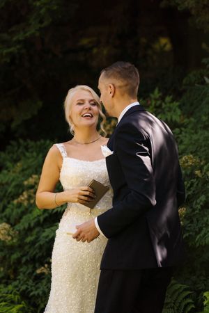 Bride in white beaded gown laughs joyfully with groom in black suit during outdoor wedding ceremony.