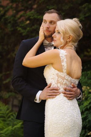 A bride in a lace gown and groom in a black tuxedo share a tender embrace outdoors surrounded by lush greenery.