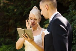 Emotional bride reads wedding vows from a small book while groom listens during outdoor ceremony.