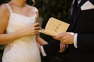 Bride in sequin gown and groom in tuxedo exchanging vows, holding gold wedding ceremony booklets outdoors.