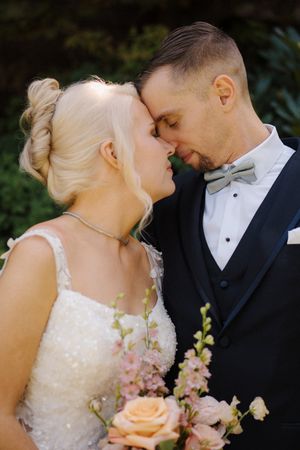 Bride and groom share romantic forehead touch in garden, bride holds pink and peach rose bouquet.