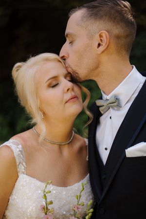 Groom in black tuxedo with sage bow tie kisses blonde bride in sparkly white dress outdoors.