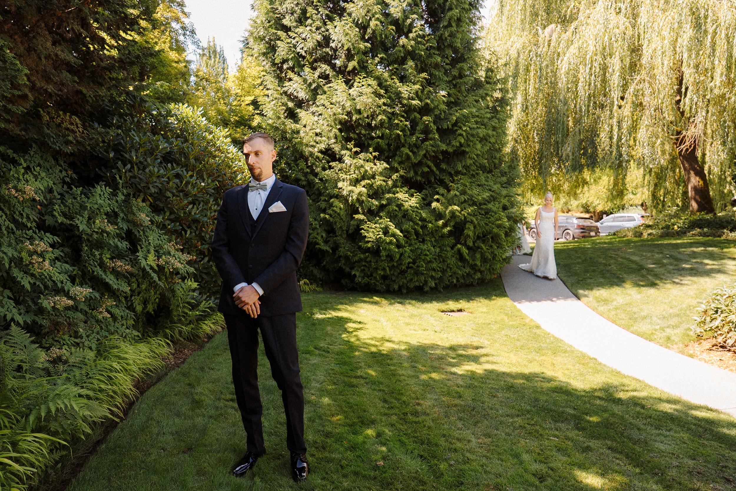 Groom in black tuxedo standing on lush green lawn surrounded by tall trees and willow at outdoor wedding venue.