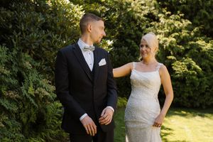 Bride in beaded gown and groom in black tuxedo share a romantic moment outdoors surrounded by lush green trees.