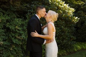 A bride and groom share a romantic kiss in front of lush green hedges during their outdoor wedding ceremony.