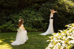 Bride and groom share a tender first look moment in a lush garden, with a flower girl watching nearby.