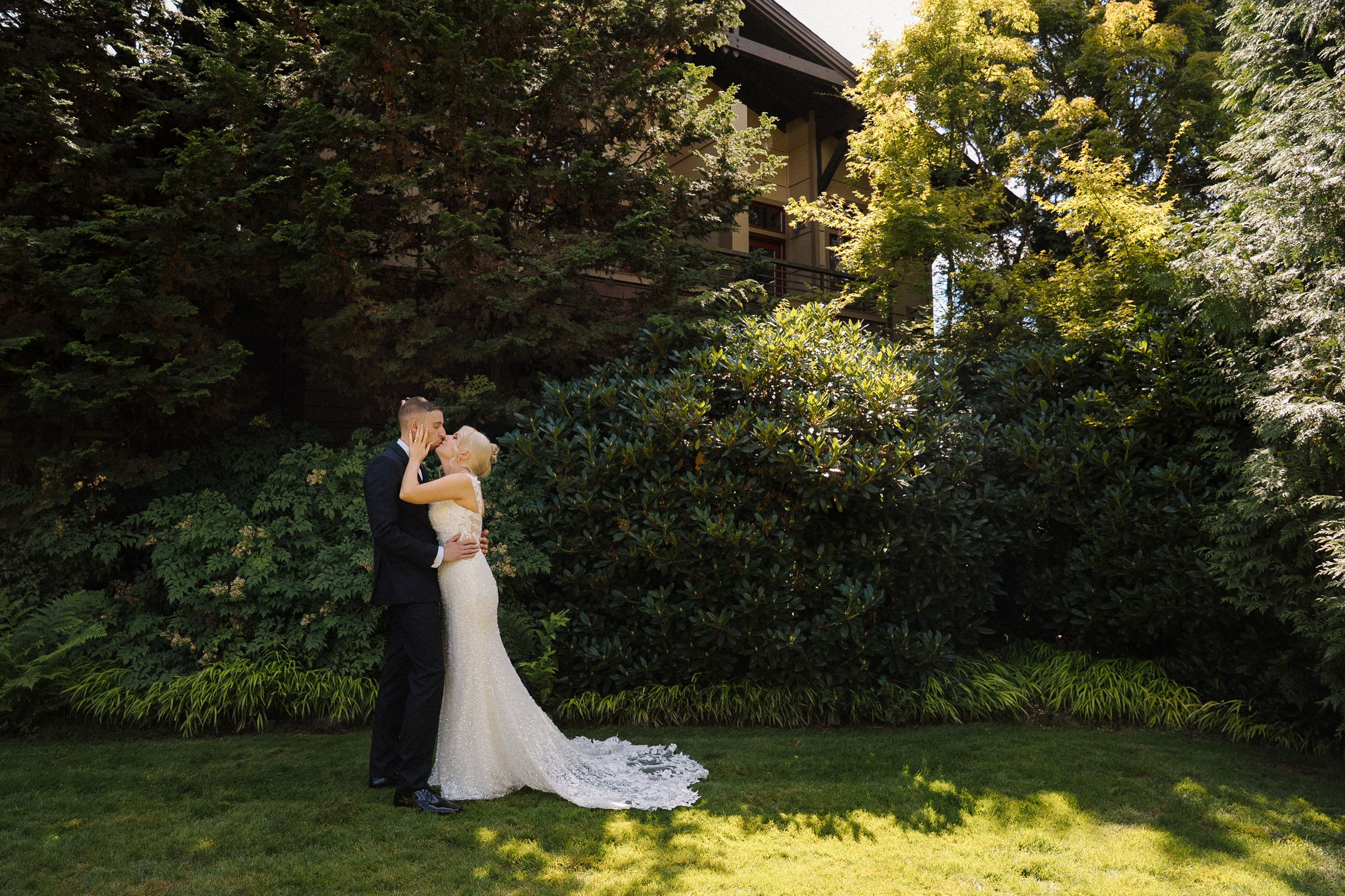 Bride and groom embrace on lush green lawn with tall trees and elegant house in background on wedding day.