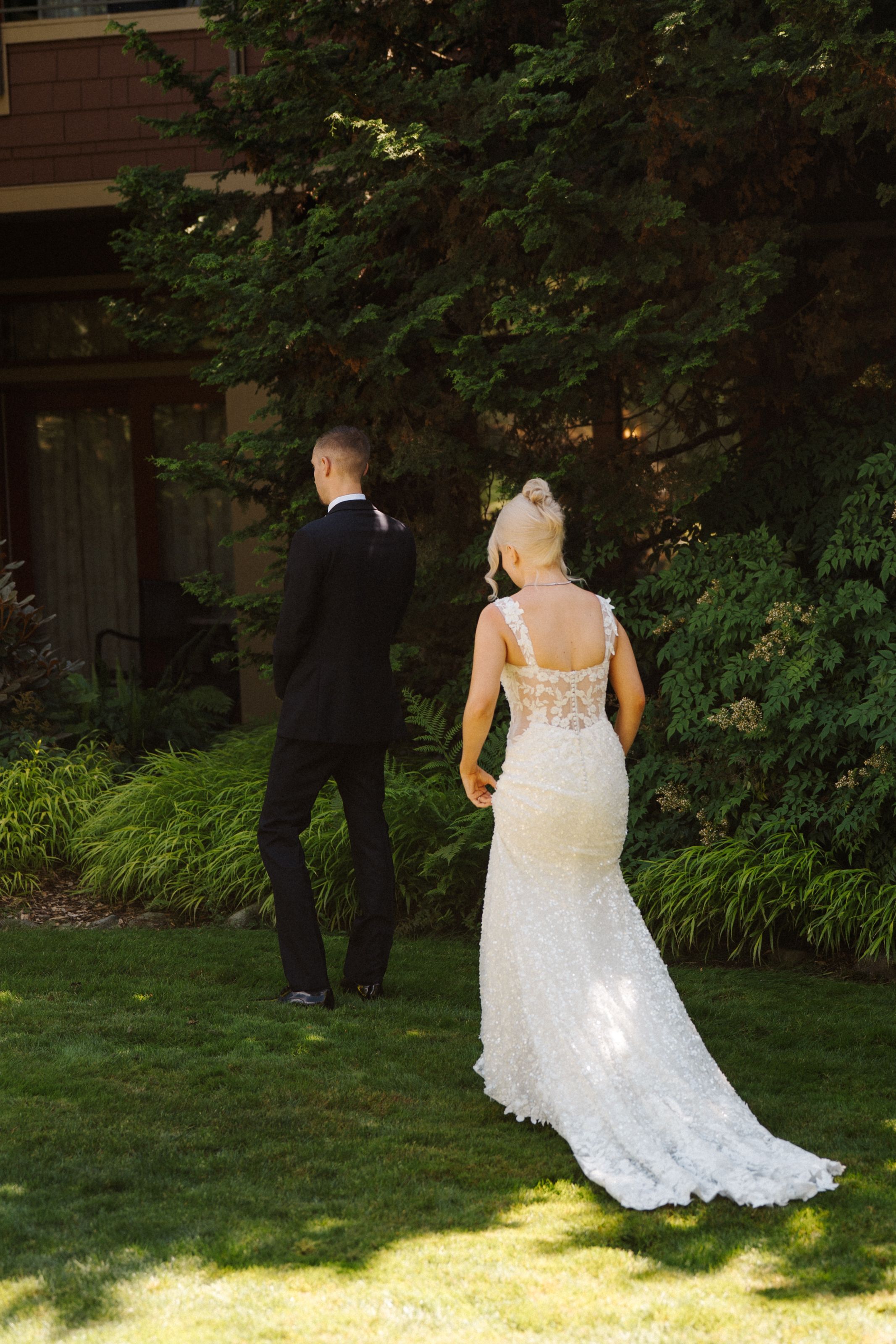 Bride in lace gown and groom in black tuxedo stand on green lawn before their first look at outdoor wedding.