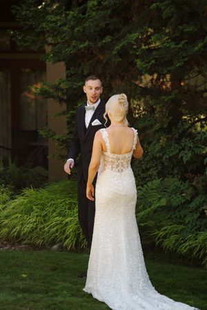 Bride in beaded gown and groom in tuxedo share a first look moment outdoors surrounded by lush green trees.