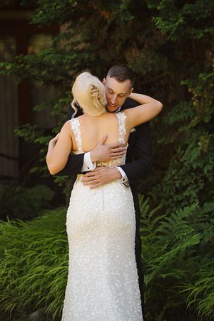 Bride and groom share a romantic embrace outdoors, her beaded white gown and his black tuxedo framed by lush green foliage.