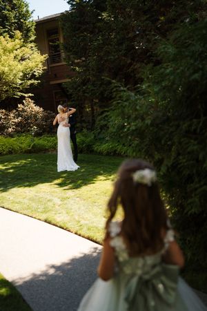 Flower girl in sage dress walks toward bride in white lace gown standing on sunlit garden lawn near trees.