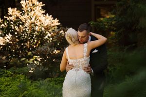 A bride and groom share a tender embrace outdoors, surrounded by lush greenery and blooming white flowers.