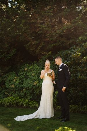 Bride in lace gown reads wedding vows from a book while groom in black tuxedo listens in a lush garden.