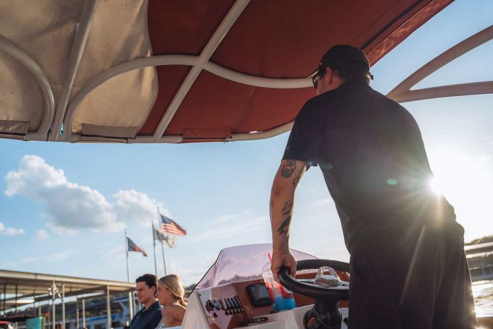 Man with tattoos steering a boat under a canopy on a sunny day with an American flag in the background.