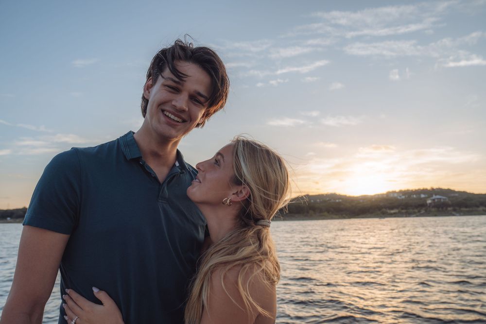Couple embracing at sunset by the water, smiling romantically in golden evening light.