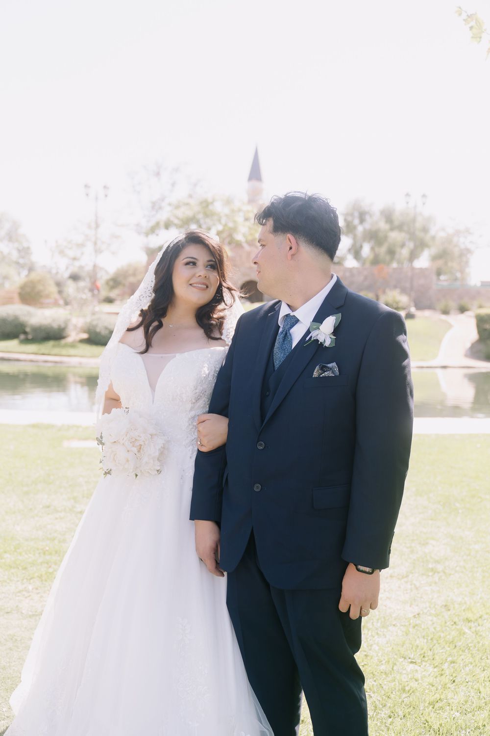 Bride in white gown and groom in navy suit pose together outdoors near a pond on their wedding day.
