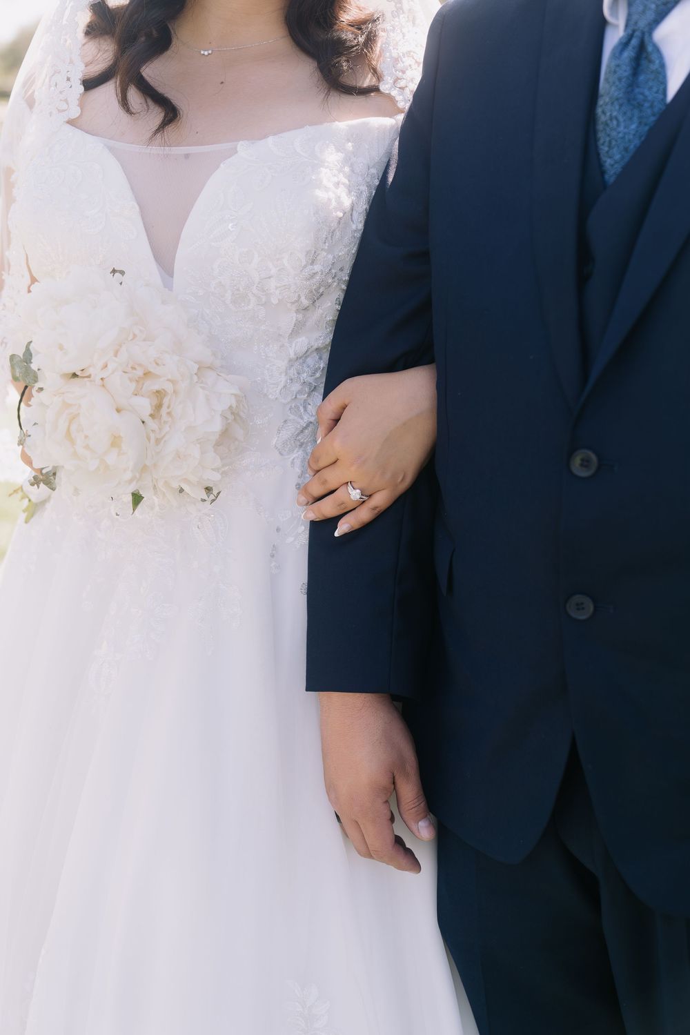 Bride in lace wedding gown holding white bouquet links arms with groom in navy suit, showing diamond ring.