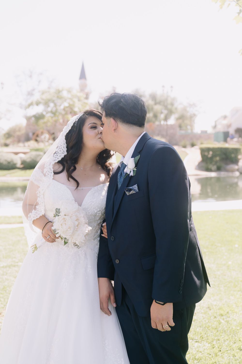 Bride and groom share a kiss outdoors, bride in lace gown with veil holding white bouquet, groom in navy suit.