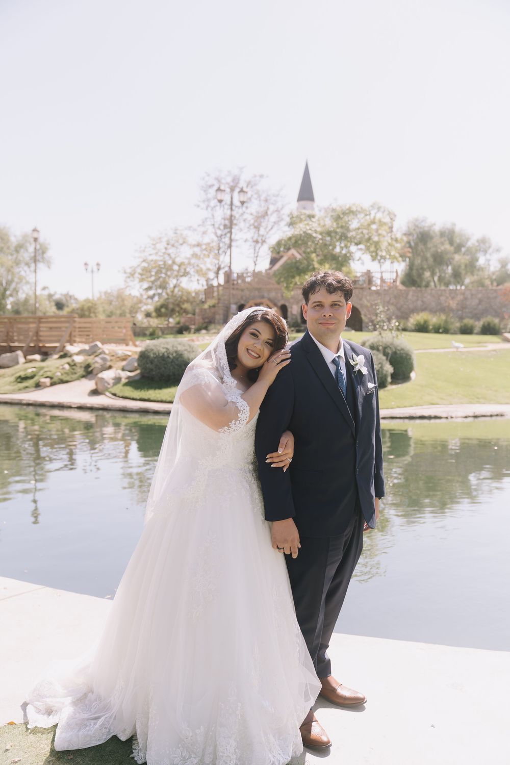 Bride in white lace gown leans on groom in navy suit by a pond at an outdoor wedding venue.
