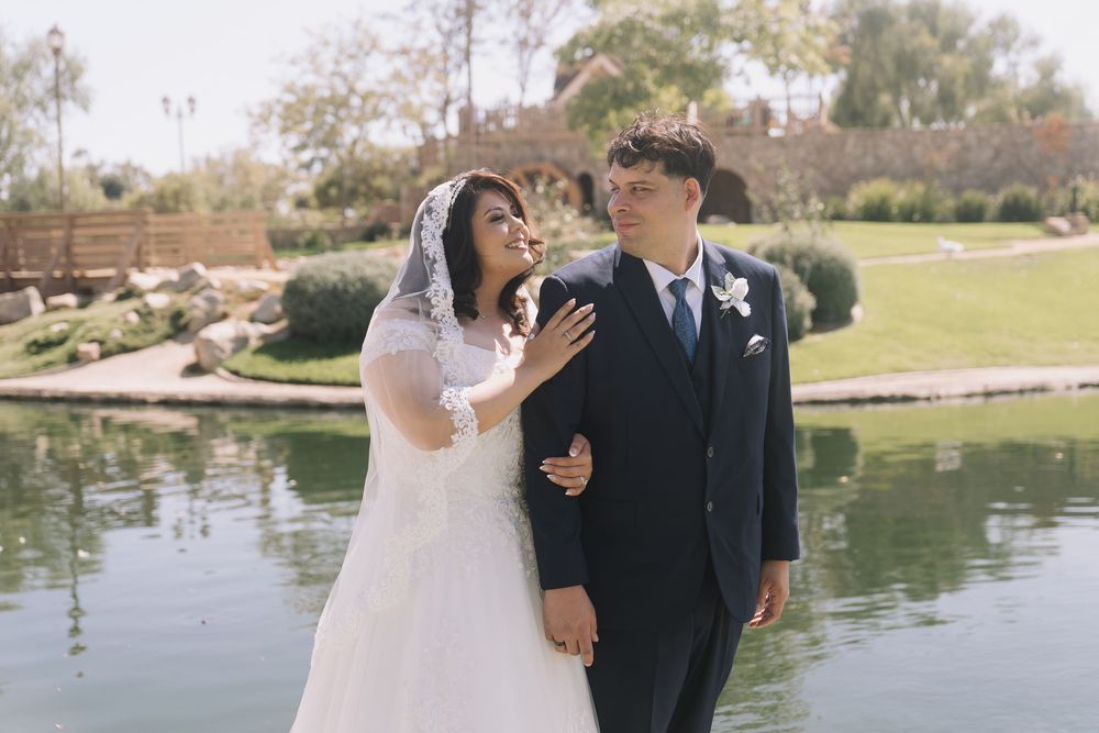 A bride in a white gown and veil stands with a groom in a dark suit by a serene pond surrounded by lush greenery.