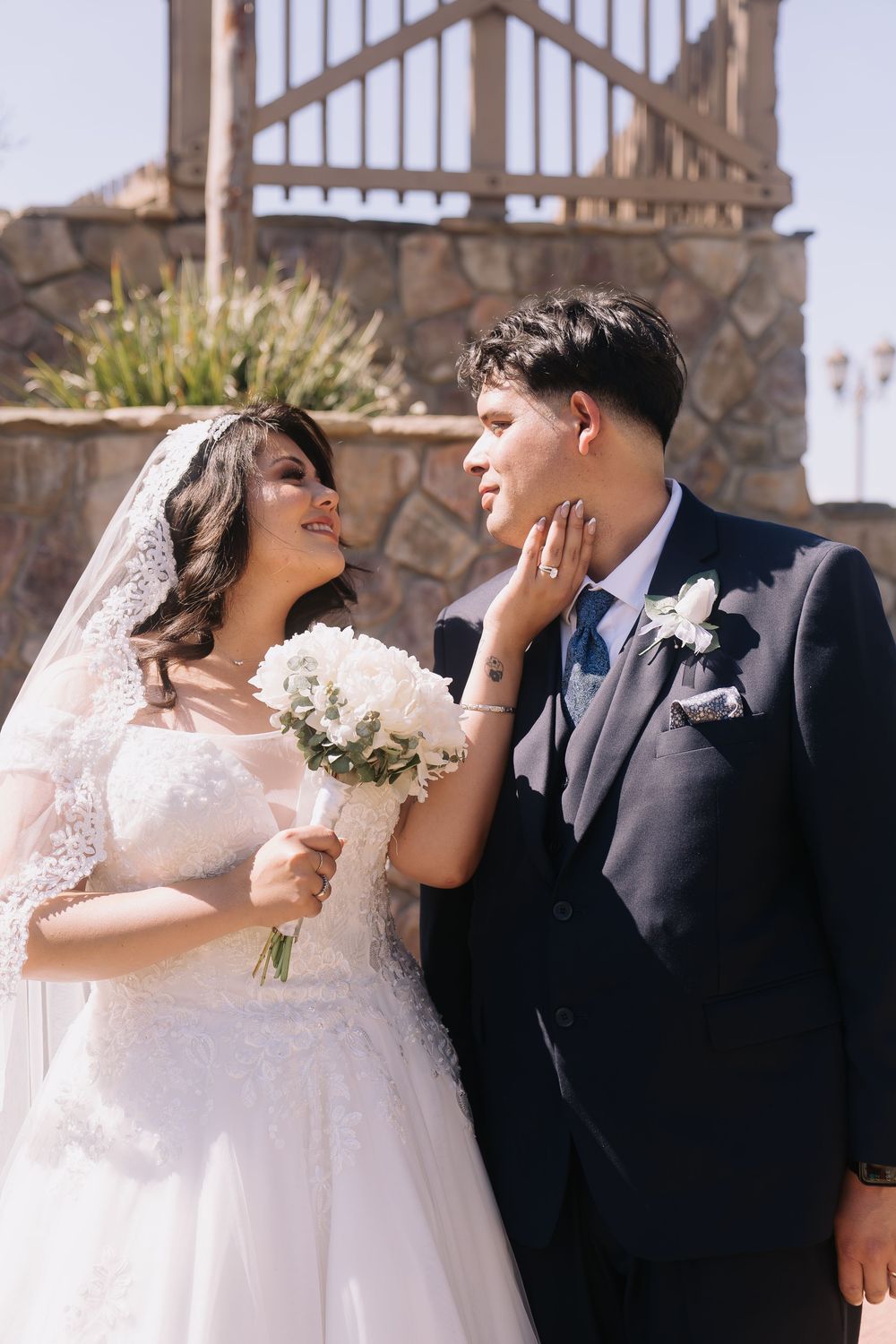 Bride in lace gown holds white bouquet, gently touching grooms face as they gaze at each other outdoors.