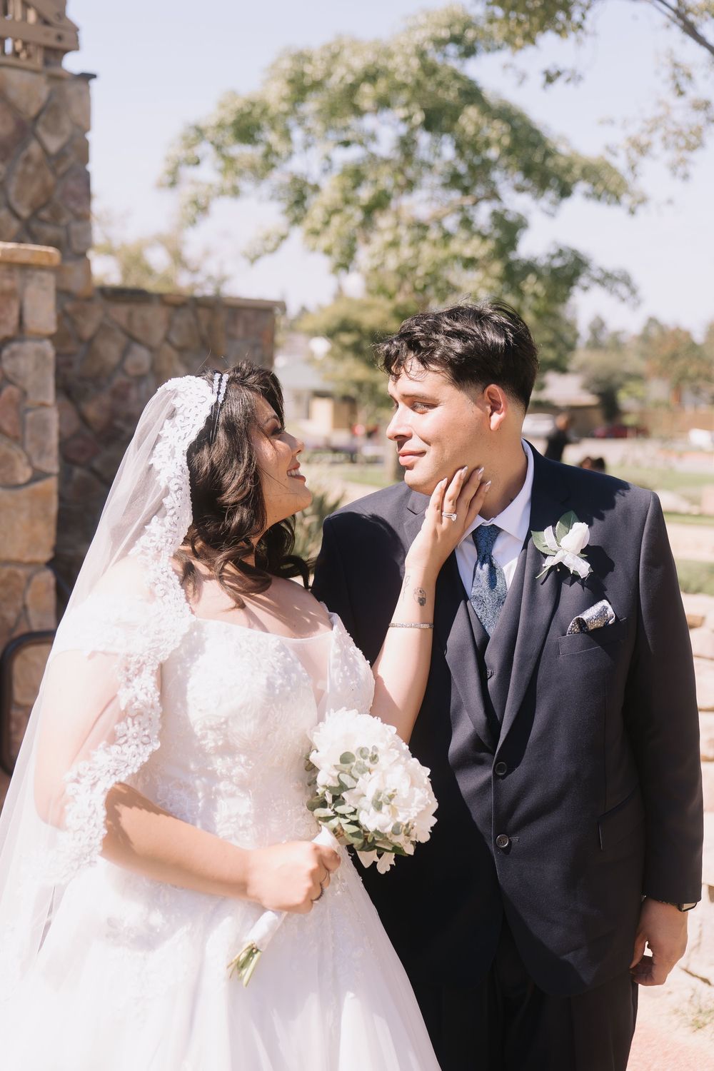 Bride in lace wedding dress tenderly touches grooms face as they gaze at each other near stone wall outdoors.