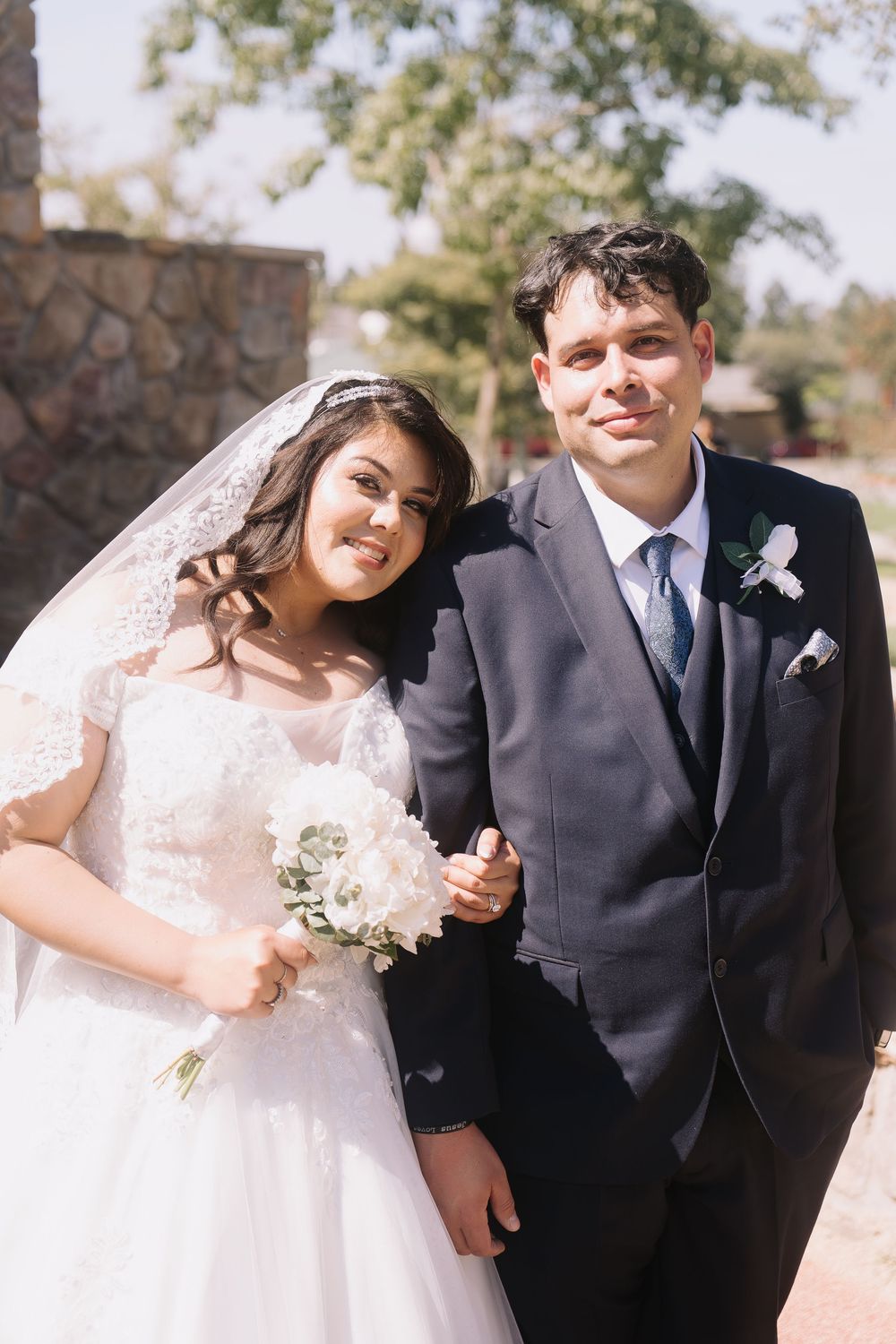 Bride in lace veil and groom in navy suit pose arm-in-arm holding white bouquet at outdoor wedding venue.