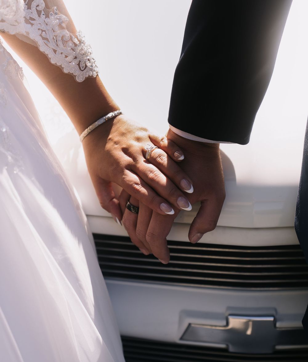 Close-up of newlyweds holding hands showing wedding rings beside a white Chevrolet vehicle.
