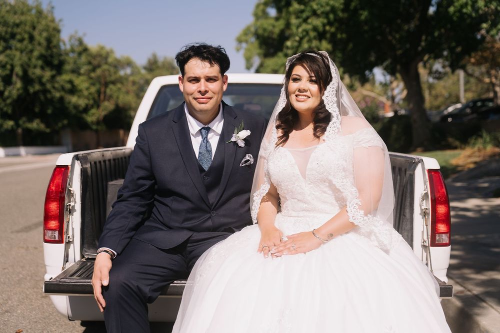 Bride in white gown and veil sits with groom in navy suit in truck bed on sunny day.