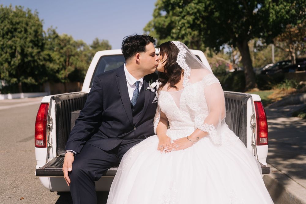 Bride and groom share a kiss while seated on the tailgate of a white pickup truck outdoors.