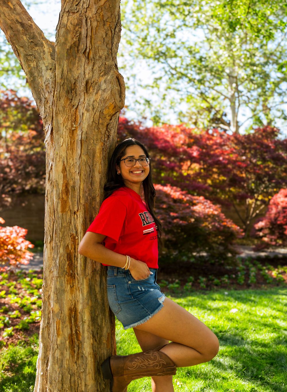 Young woman in red college shirt and denim shorts smiling while leaning against a tree in a colorful garden.