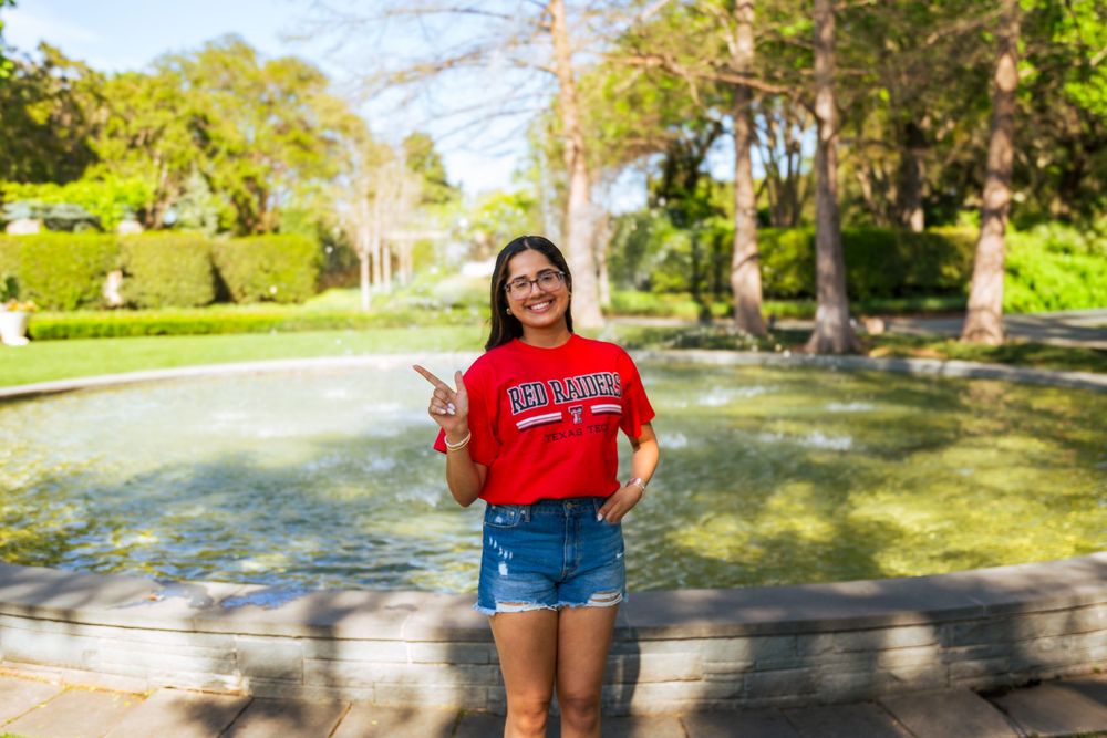 A smiling young woman in a red college shirt and denim shorts poses by a fountain in a sunny park.