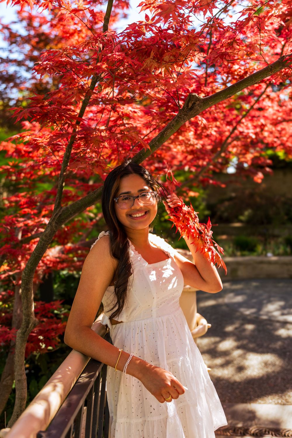 Smiling young woman in white dress posing under vibrant red Japanese maple tree in a garden.