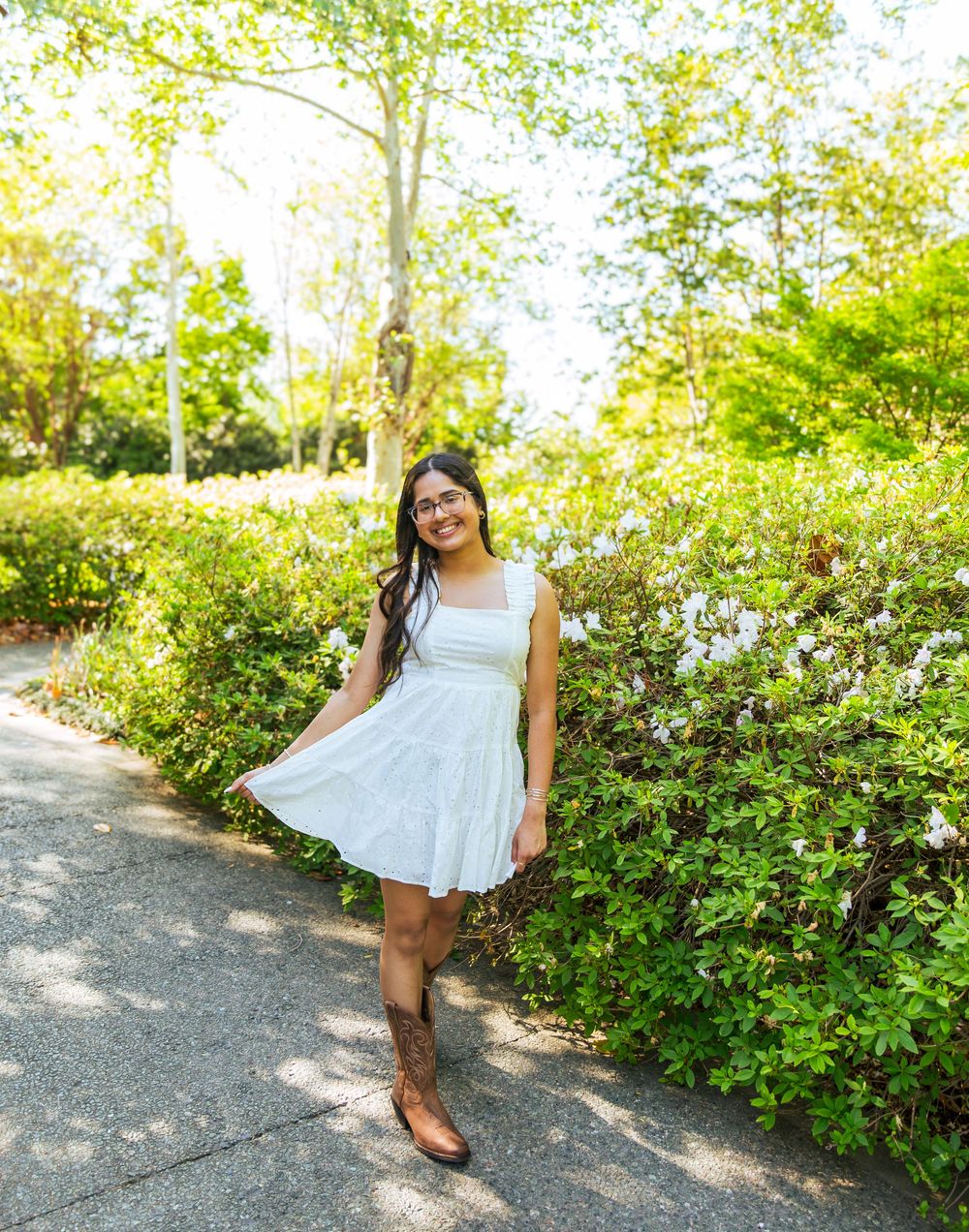 Young woman in white dress and cowboy boots smiling on a garden path surrounded by lush green shrubs and trees.