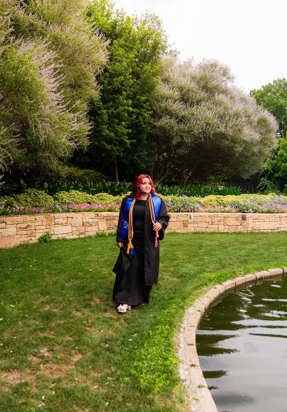 Graduate in black gown with colorful stole stands by a garden pond with lush green trees in background.