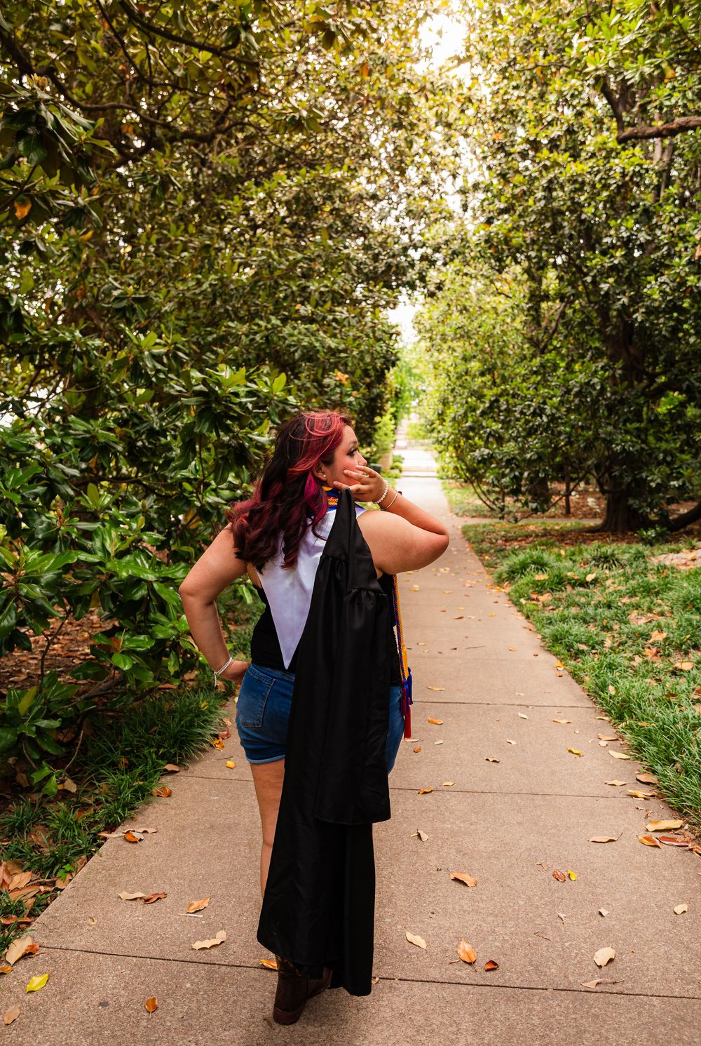 Woman with red hair walks down a tree-lined path holding a black graduation gown on a sunny day.
