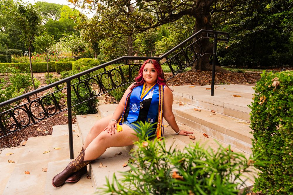 Woman with red hair in blue outfit sits on stone steps surrounded by lush greenery in a park setting.