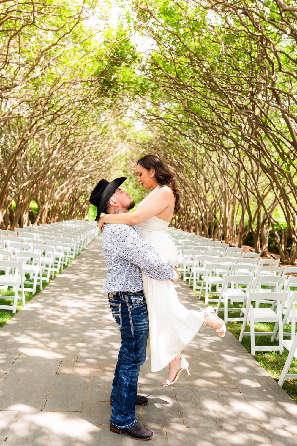 Groom lifting bride in white dress at outdoor wedding venue with tree canopy and white chairs.