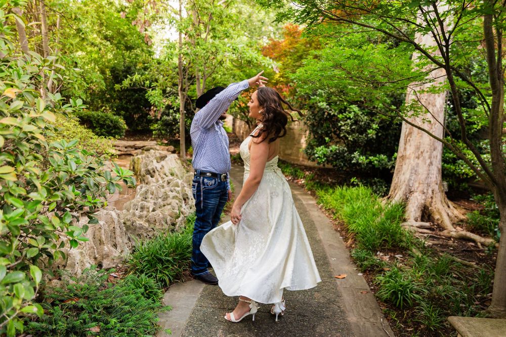 A bride in a white dress twirls with her groom on a garden path surrounded by lush green trees.
