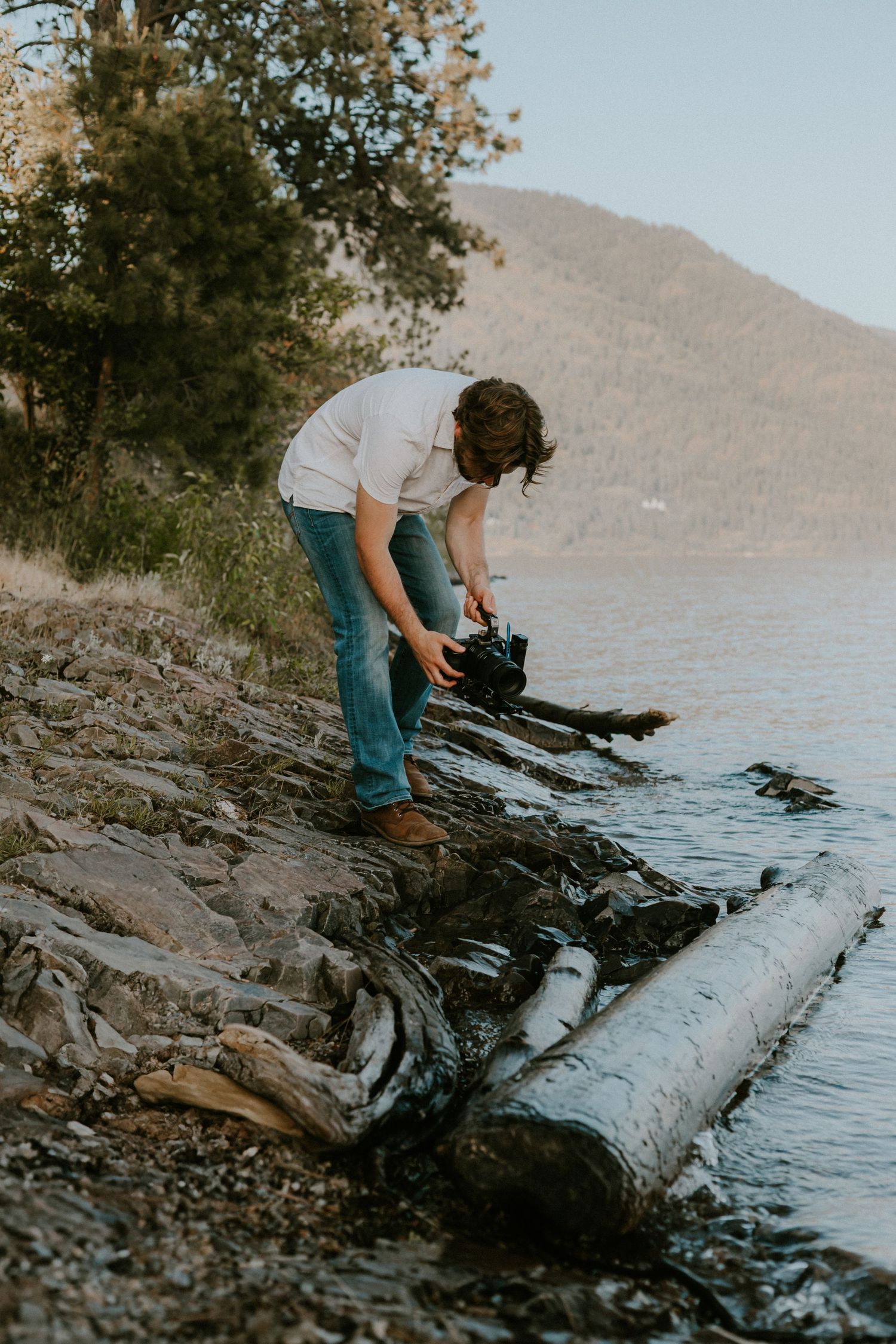 Person in white shirt and jeans photographing on rocky lakeside shore with mountains and trees in background.