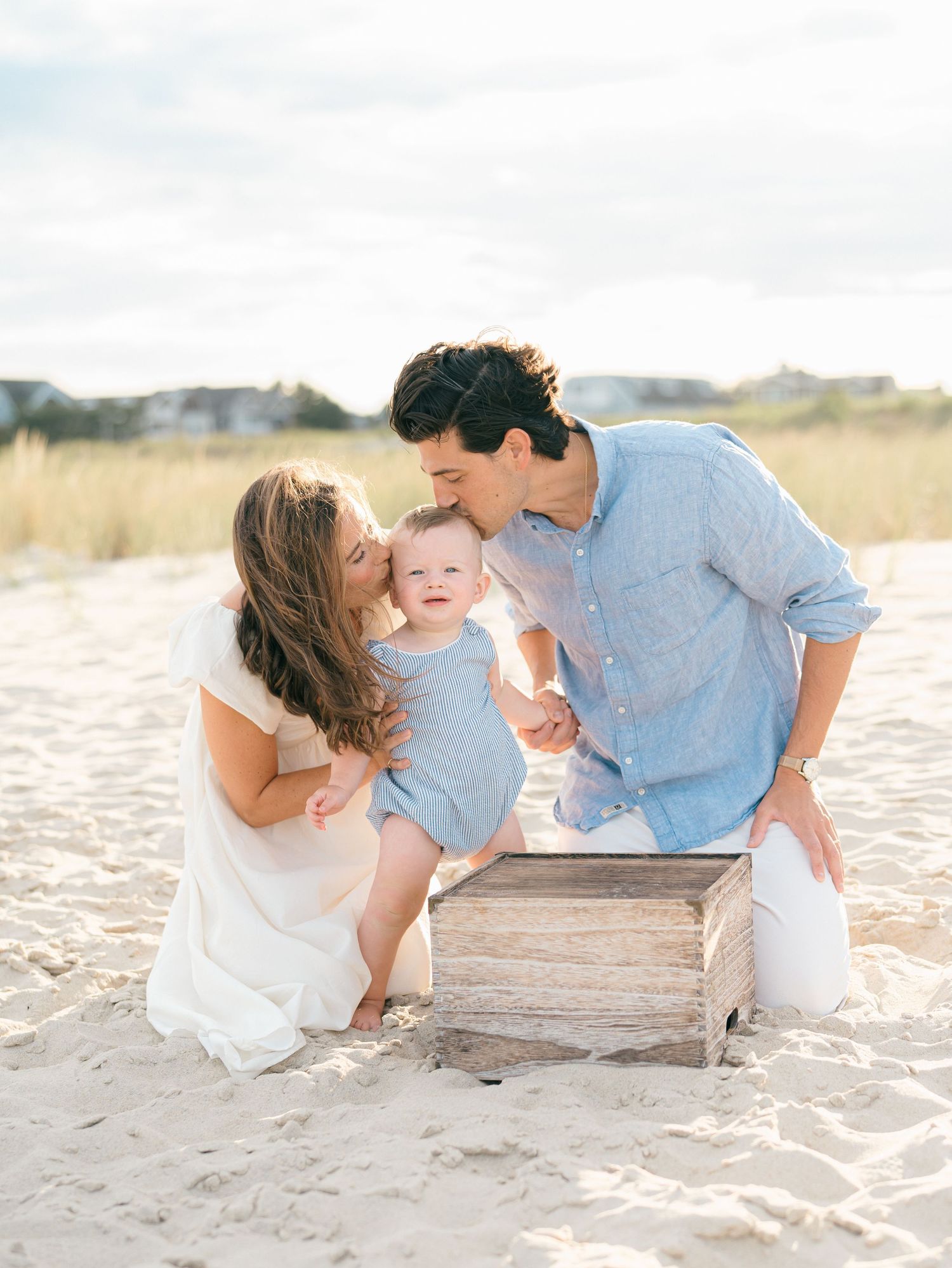 Man in blue collar shirt and wife in white dress play with their toddler on the spring lake beach.