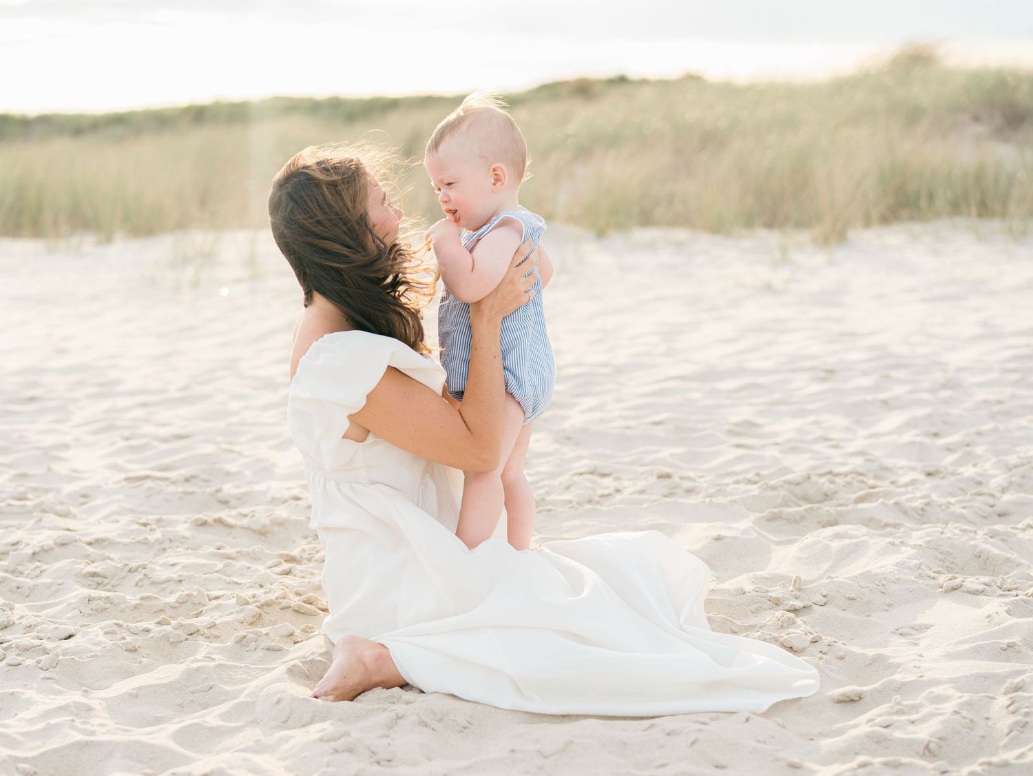 A mother holds up her toddler son in the air with spring lake sand dunes in the background.