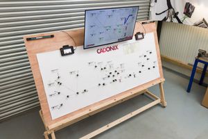 Cadonix control panel board with network diagram on wooden easel stand, featuring a monitor display in a workshop.