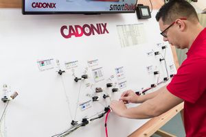 A technician in a red shirt works on a Cadonix SmartBuilder wiring diagram mounted on a white wall.