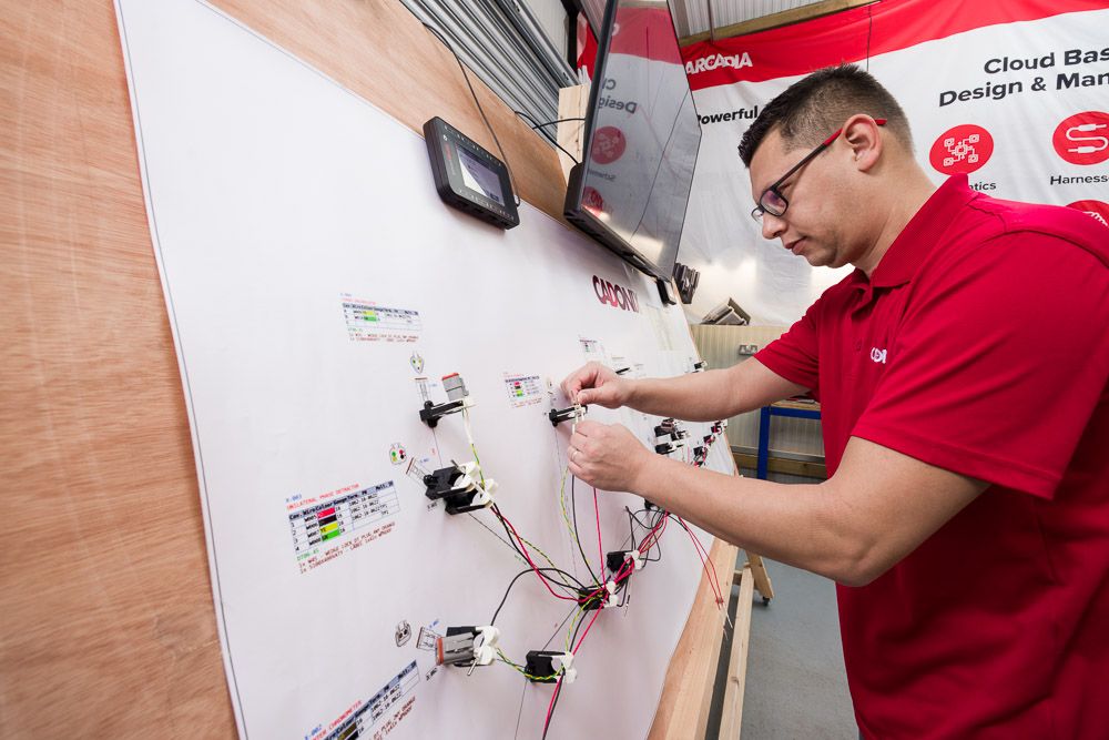 Electrician in red shirt wiring electrical components on a white board in a professional training environment.