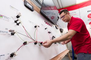 Electrician in red shirt wiring electrical components on a white board in a workshop or training facility.