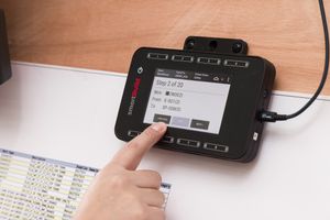 A finger touches an industrial control panel displaying step-by-step instructions, placed on a wooden surface.