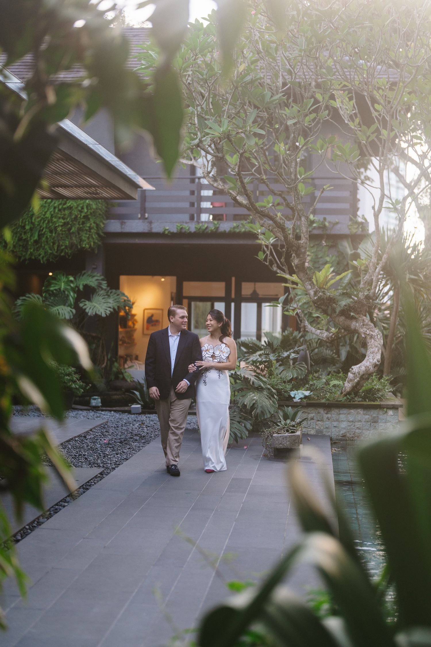 Bride and groom embracing by reflective pool before elegant building with arched windows and terracotta roof.